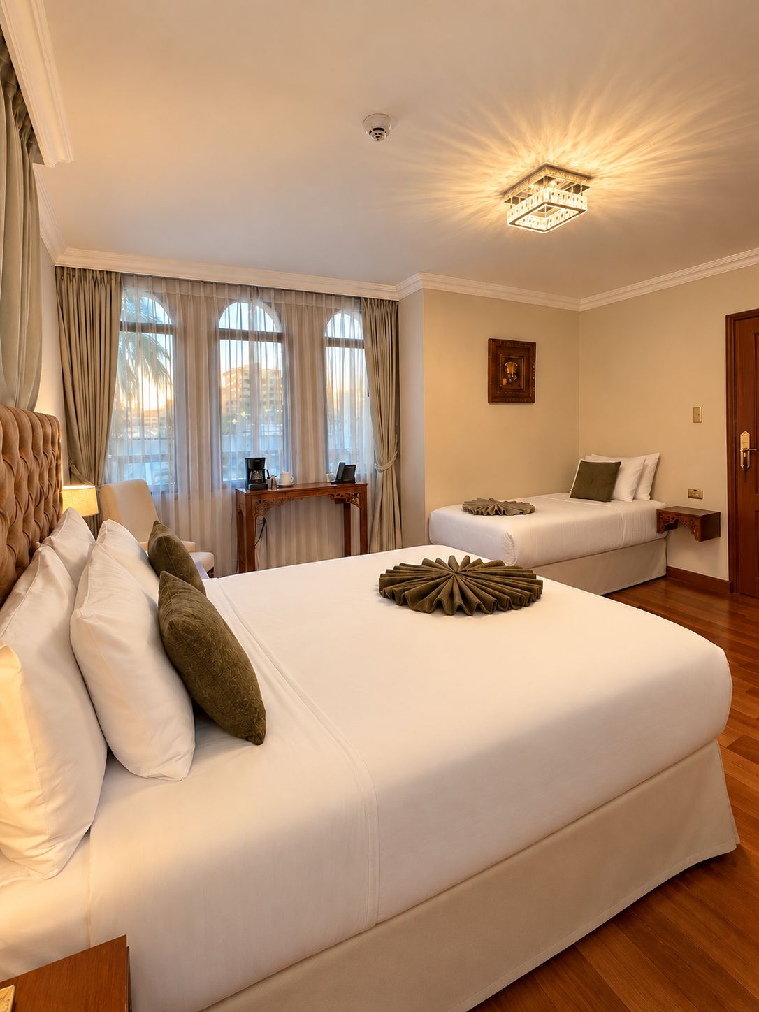 Ein Hotelzimmer mit einem großen Bett, einem kleineren Bett, einem Schreibtisch mit Stuhl und einem fenster mit Vorhängen.