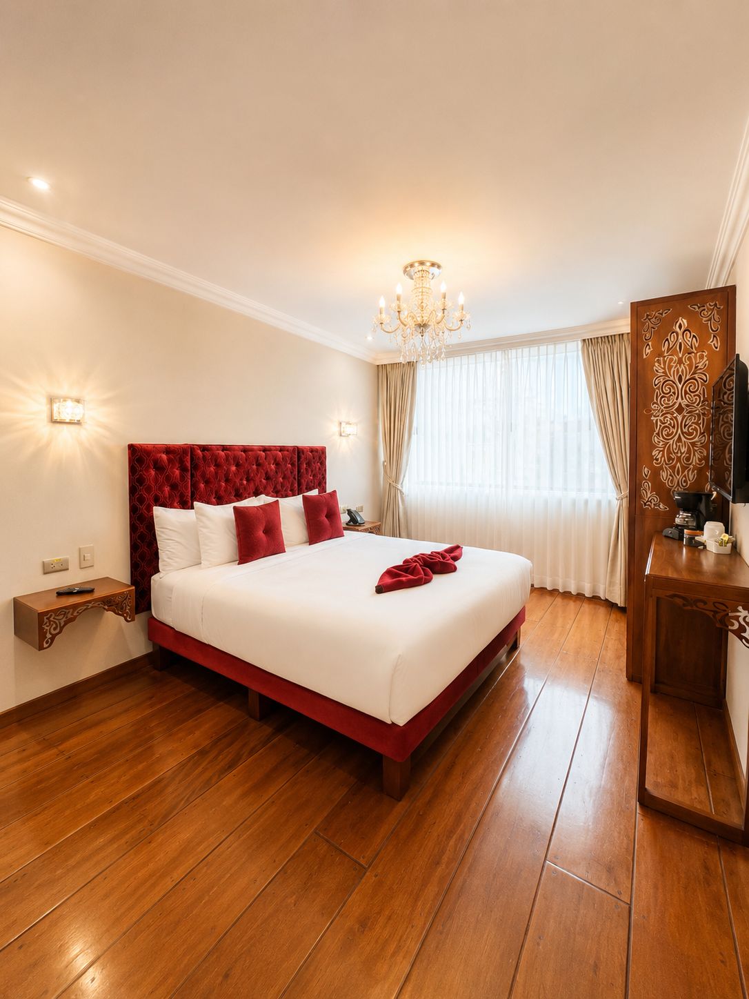 A hotel room with a large bed, wooden flooring, a chandelier, a TV, and a decorative mirror.