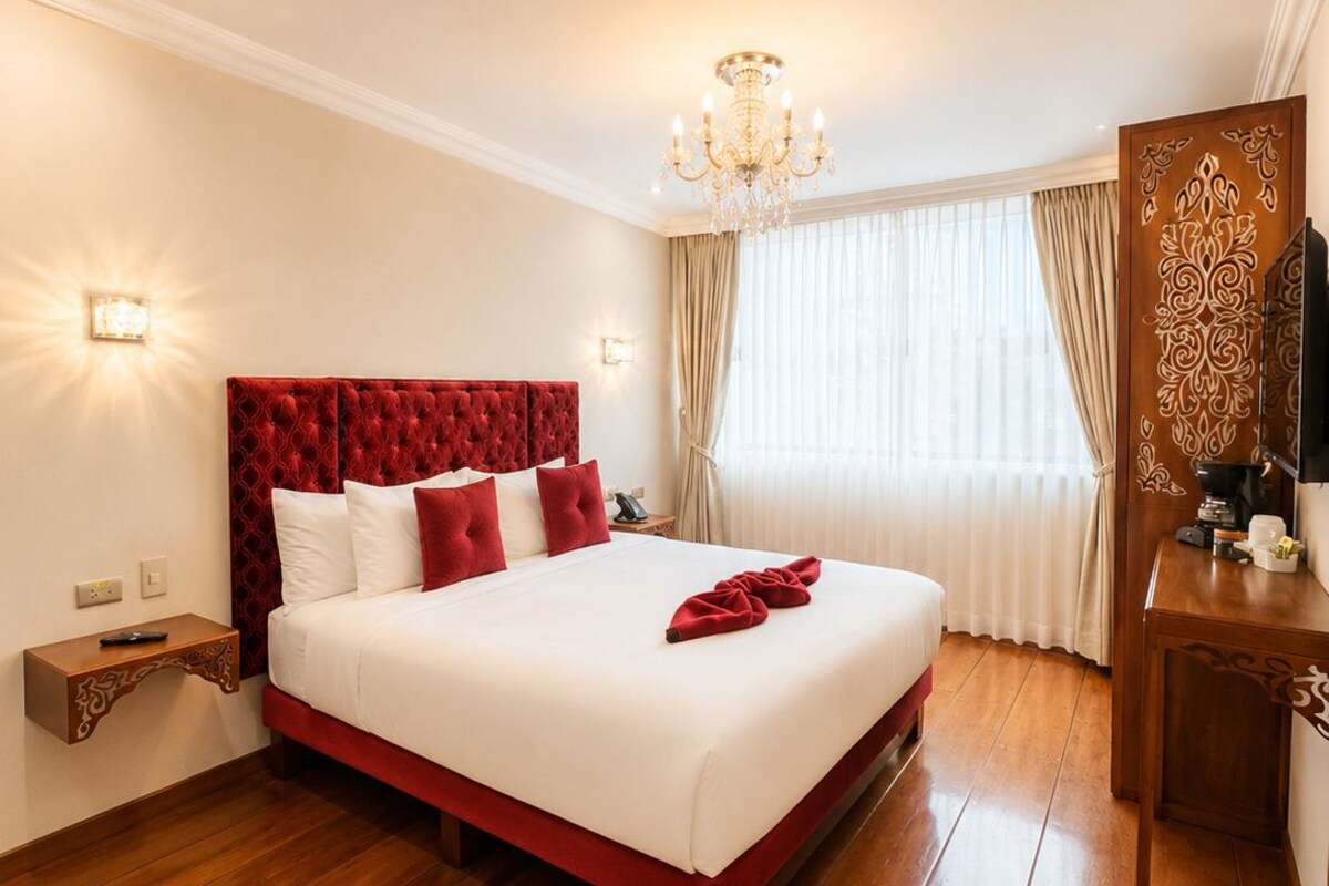 A hotel room with a large bed, wooden flooring, a chandelier, a TV, and a decorative mirror.