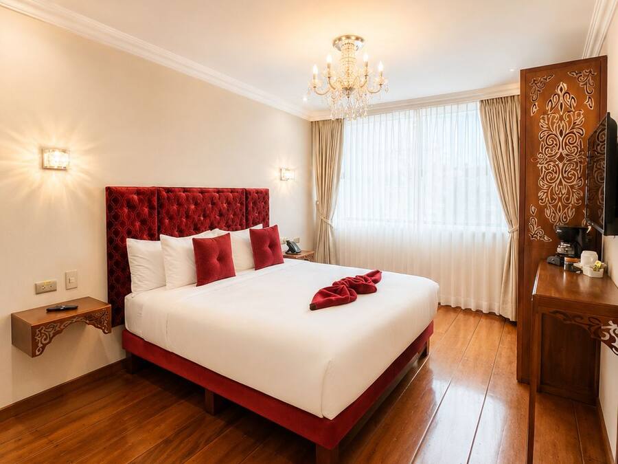 A hotel room with a large bed, wooden flooring, a chandelier, a TV, and a decorative mirror.