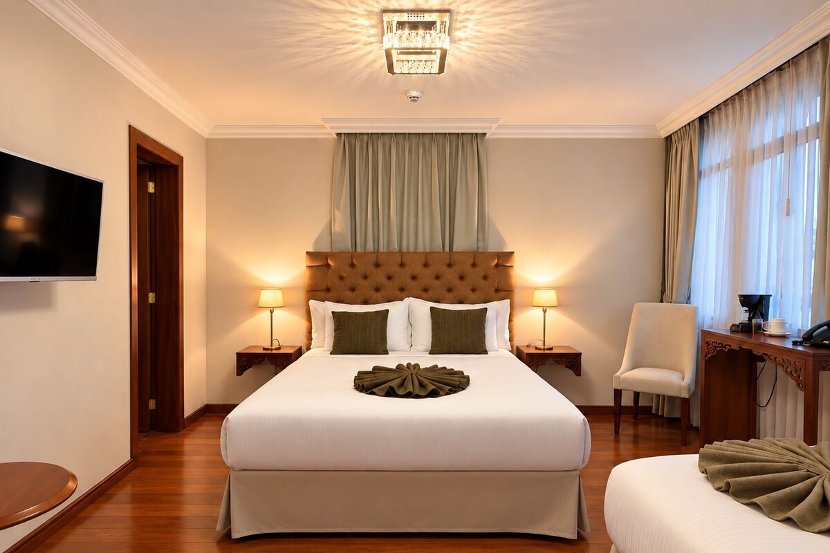 A hotel room with a large bed, two bedside tables with lamps, a flat-screen TV, a wooden desk with a chair, and a window with curtains.