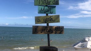 A wooden sign with text in a foreign language, a sandy beach, and a clear blue sky. - Branca Tropical Brasil Residencial (Porto Seguro)