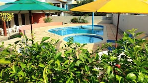 A swimming pool with two umbrellas, surrounded by greenery and a building in the background. - Branca Tropical Brasil Residencial (Porto Seguro)