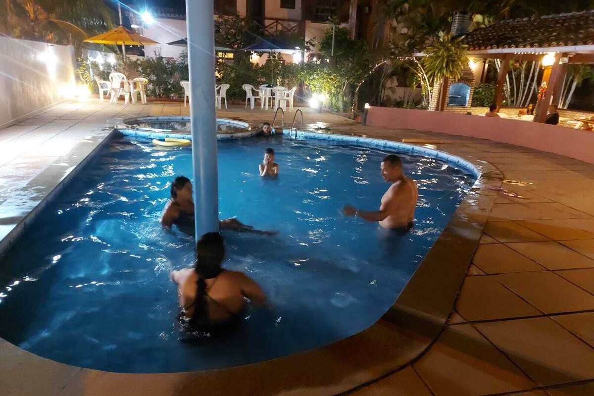 Ein Schwimmbecken mit drei Personen im Wasser, umgeben von Sitzgelegenheiten im Freien und Palmen.