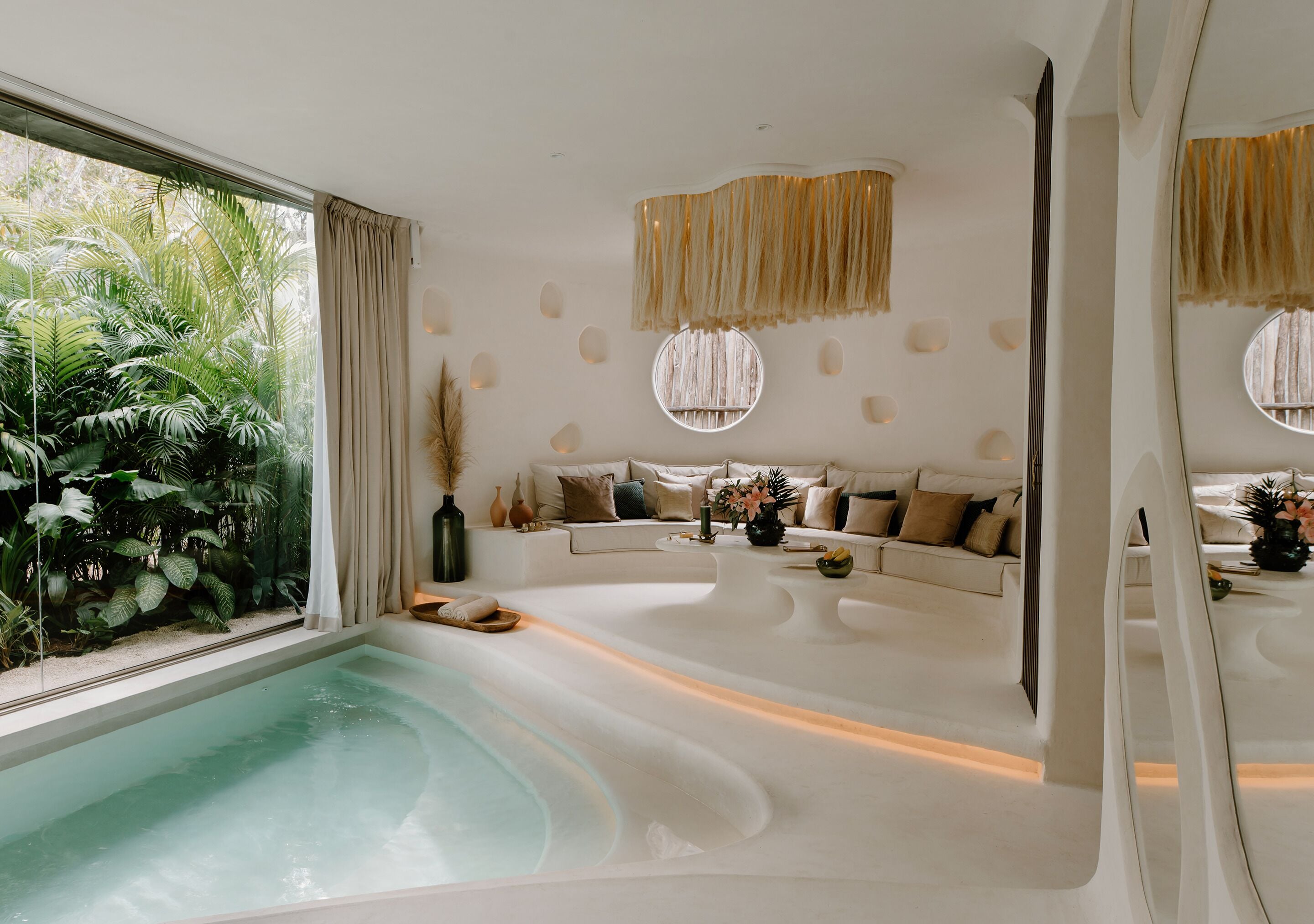 A modern spa room with a large bathtub, a sofa, and a view of lush greenery.