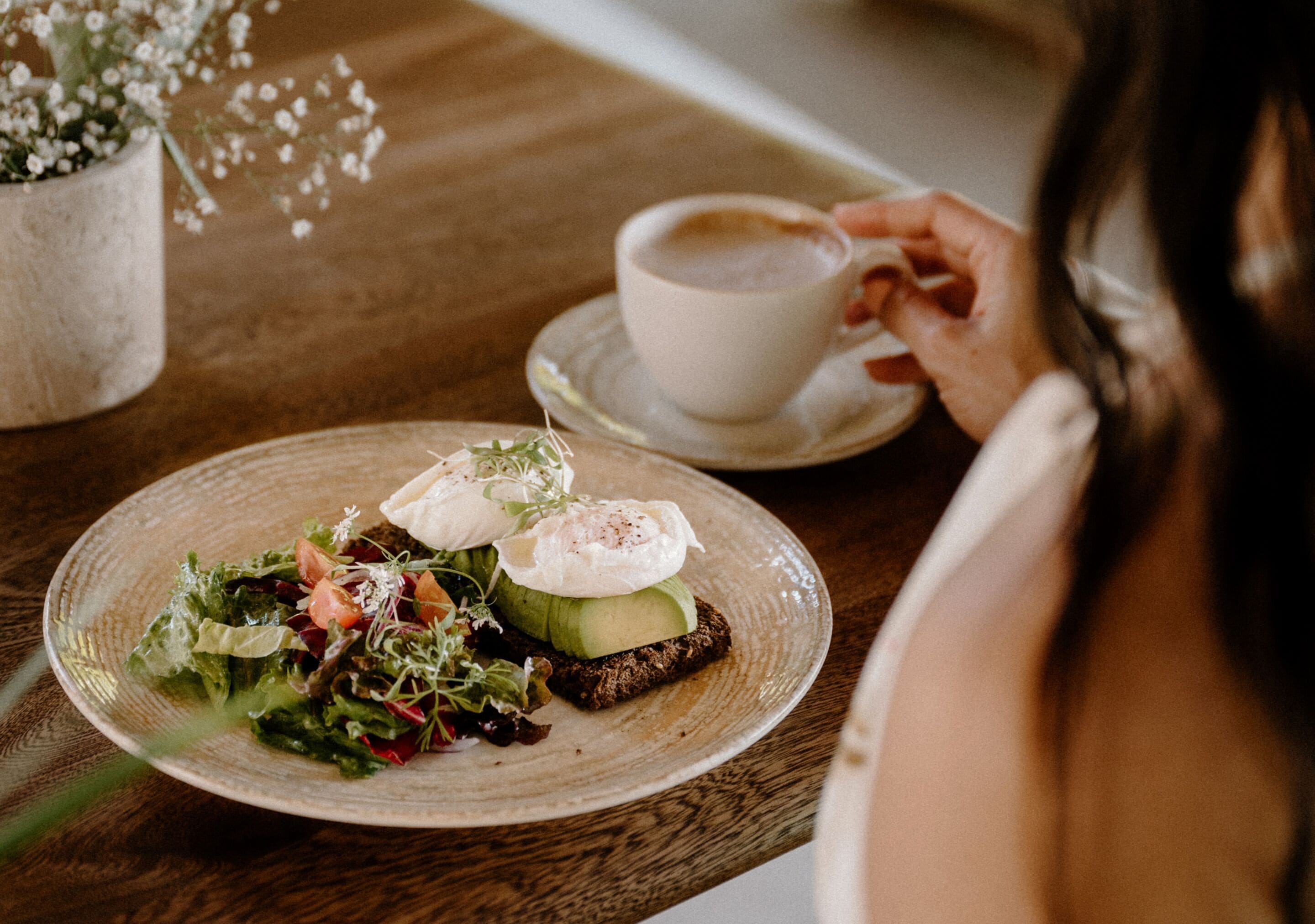 Ein Teller mit Essen, darunter ein Salat und ein pochiertes Ei auf Toast, eine Tasse Kaffee und ein kleiner Topfpflanze auf einem Holztisch.