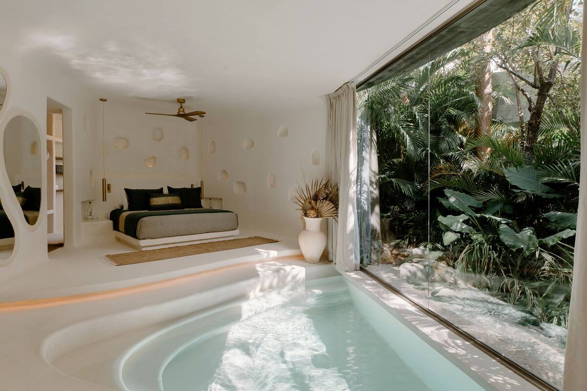 Un dormitorio moderno con un ventanal grande, una bañera integrada y vistas a vegetación exuberante.