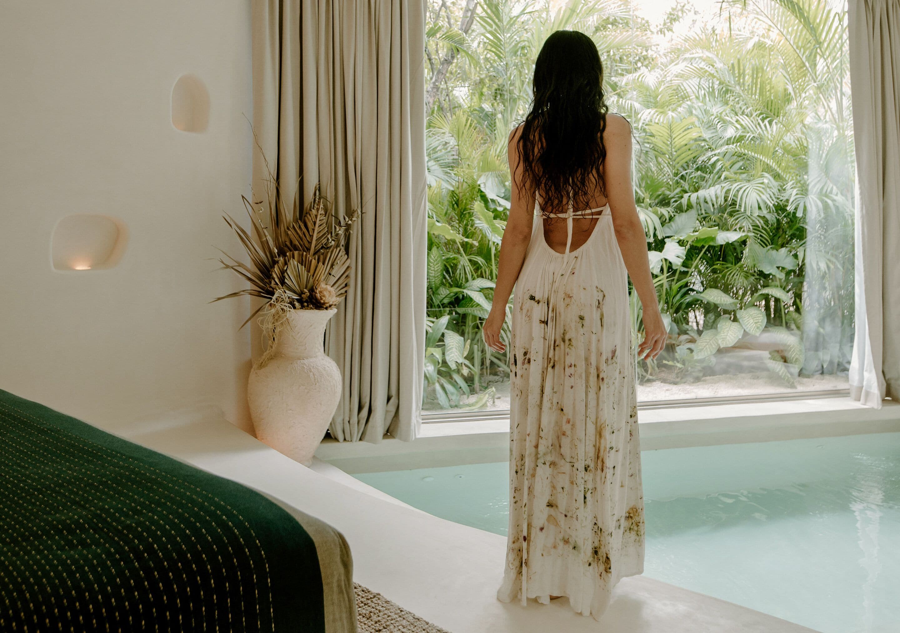 A woman in a white dress standing by a window with a view of greenery.