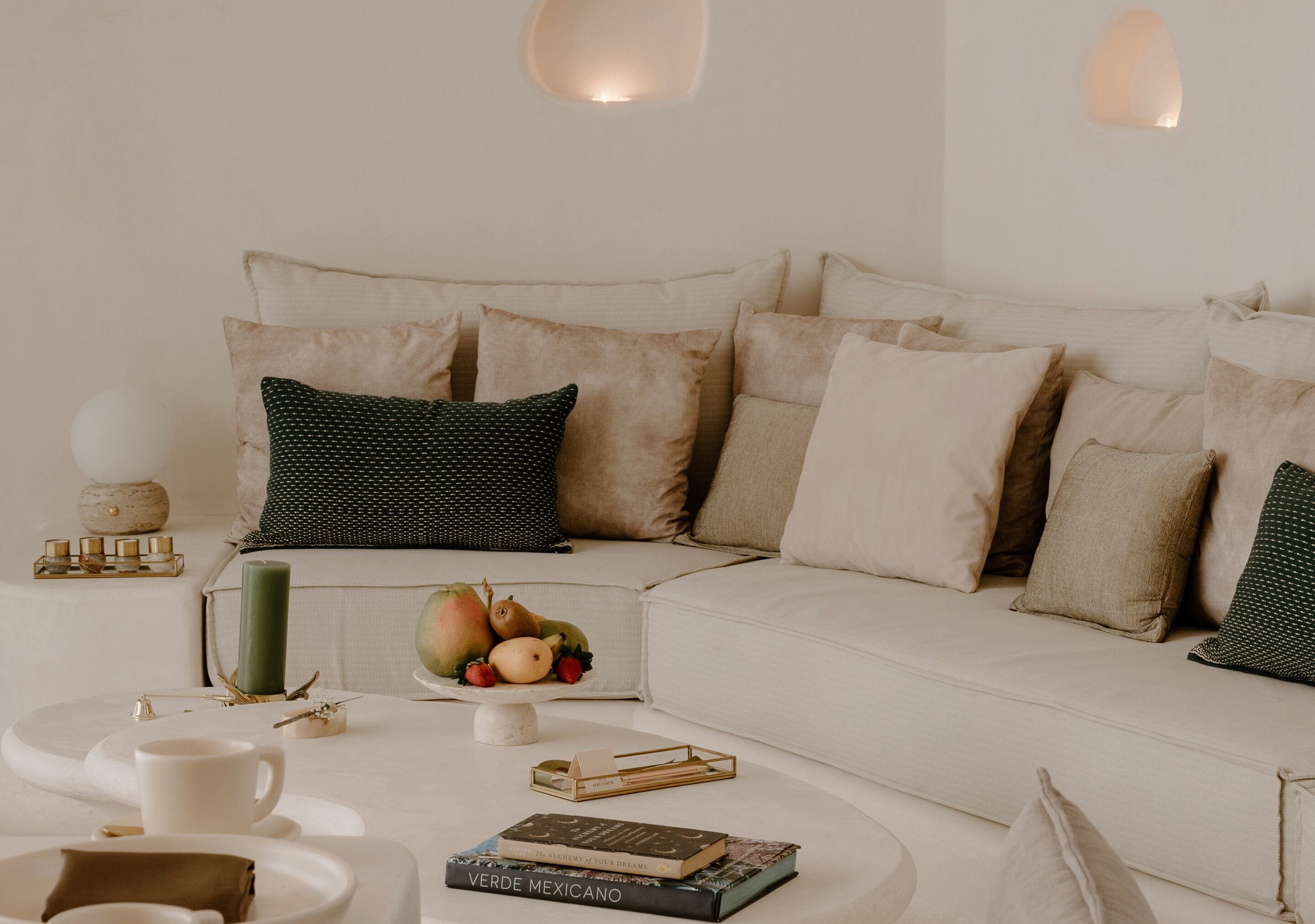 Un salon moderne avec un canapé blanc, de nombreux coussins, une table basse avec des livres et une bougie, et un panier de fruits.