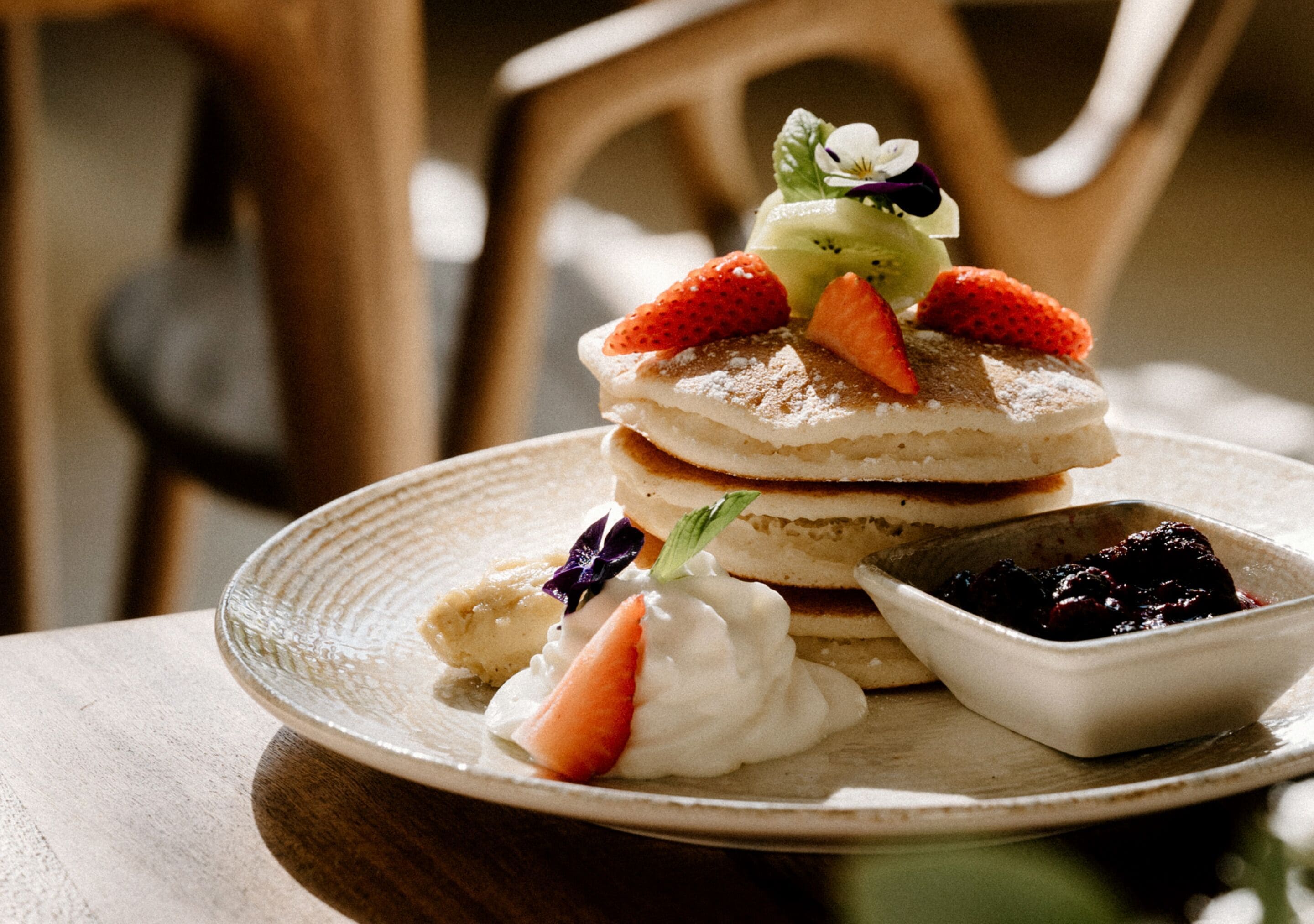 A stack of pancakes with fruit on top, a small bowl of berries, and a dollop of whipped cream on a plate.