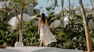 Une femme vêtue d’une robe blanche marche sur un sentier bordé d’une végétation luxuriante et entouré de structures sphériques blanches.