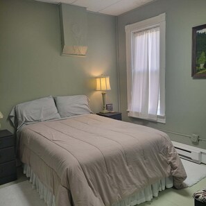 Un dormitorio con cama, mesita de noche, lámpara, ventana con cortinas y un cuadro en la pared.