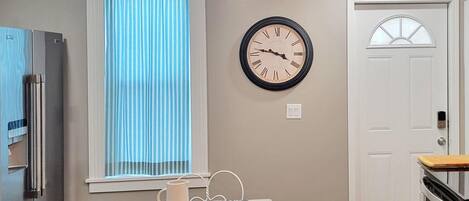 A kitchen with a clock, a window with blinds, a refrigerator, and a door.
