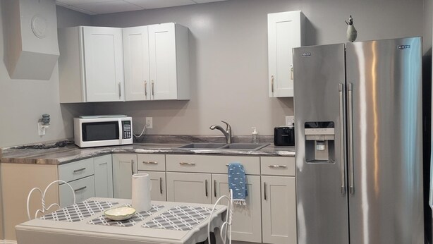 A modern kitchen with white cabinets, a stainless steel refrigerator, a microwave, and a small dining area with a table and chairs. - Warm northwoods beach-style apartment walking distance to OreBeGone & Mesabi Trl (Gilbert)