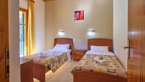 A bedroom with two wooden beds, each with a quilted bedspread and pillows. There are bedside tables with lamps, a window with curtains, and a wall-mounted light fixture. - Theodora Apartment 2 Agios Stefanos Corfu (Corfu)