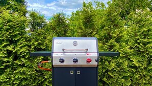 Una parrilla portátil con pantalla digital, perillas de control y un asa, frente a un fondo de follaje verde exuberante.