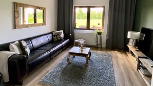 Una sala de estar con un sofá de cuero oscuro, una mesa de centro de madera, un televisor sobre un mueble de madera y una ventana con cortinas.