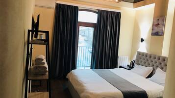 A hotel room with a bed, a shelf with towels, a window with curtains, and a painting on the wall.