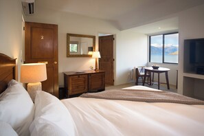 A hotel room with a bed, a desk, a chair, a TV, and a window with a view of mountains. - Hidden Lodge (Queenstown)