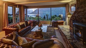 A cozy living room with a fireplace, leather sofas, a coffee table, and large windows offering a view of mountains and a lake. - Hidden Lodge (Queenstown)