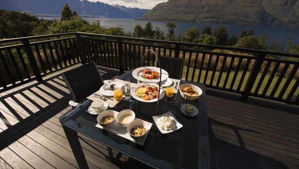 A dining table with various dishes and a glass of orange juice on a deck overlooking a lake and mountains. - Hidden Lodge (Queenstown)