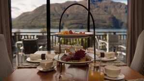 A breakfast setup with a tiered tray, plates, cups, and a glass of orange juice on a table with a view of mountains and a lake. - Hidden Lodge (Queenstown)