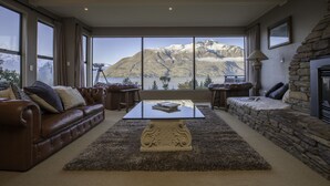 A spacious living room with a large window offering a mountain view, a leather sofa, a coffee table, and a stone fireplace. - Hidden Lodge (Queenstown)