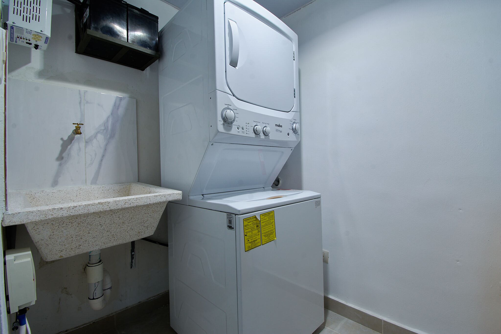 Un espace buanderie compact comprenant une machine à laver et un sèche-linge superposés, un évier avec un plan de travail et un panneau électrique au-dessus de l’évier.