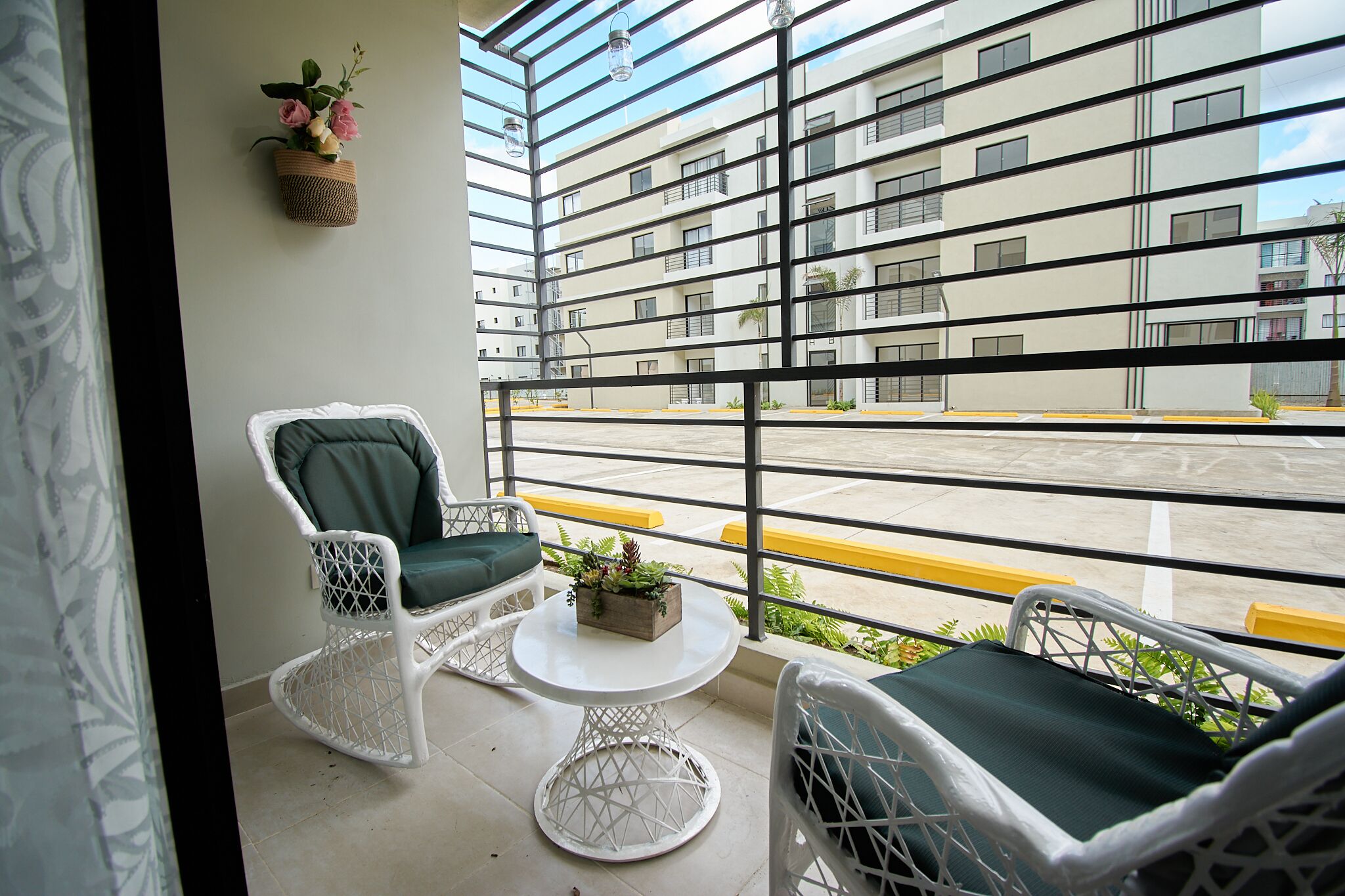 Un balcon avec des fauteuils en osier et une petite table, donnant sur un parking et des immeubles.