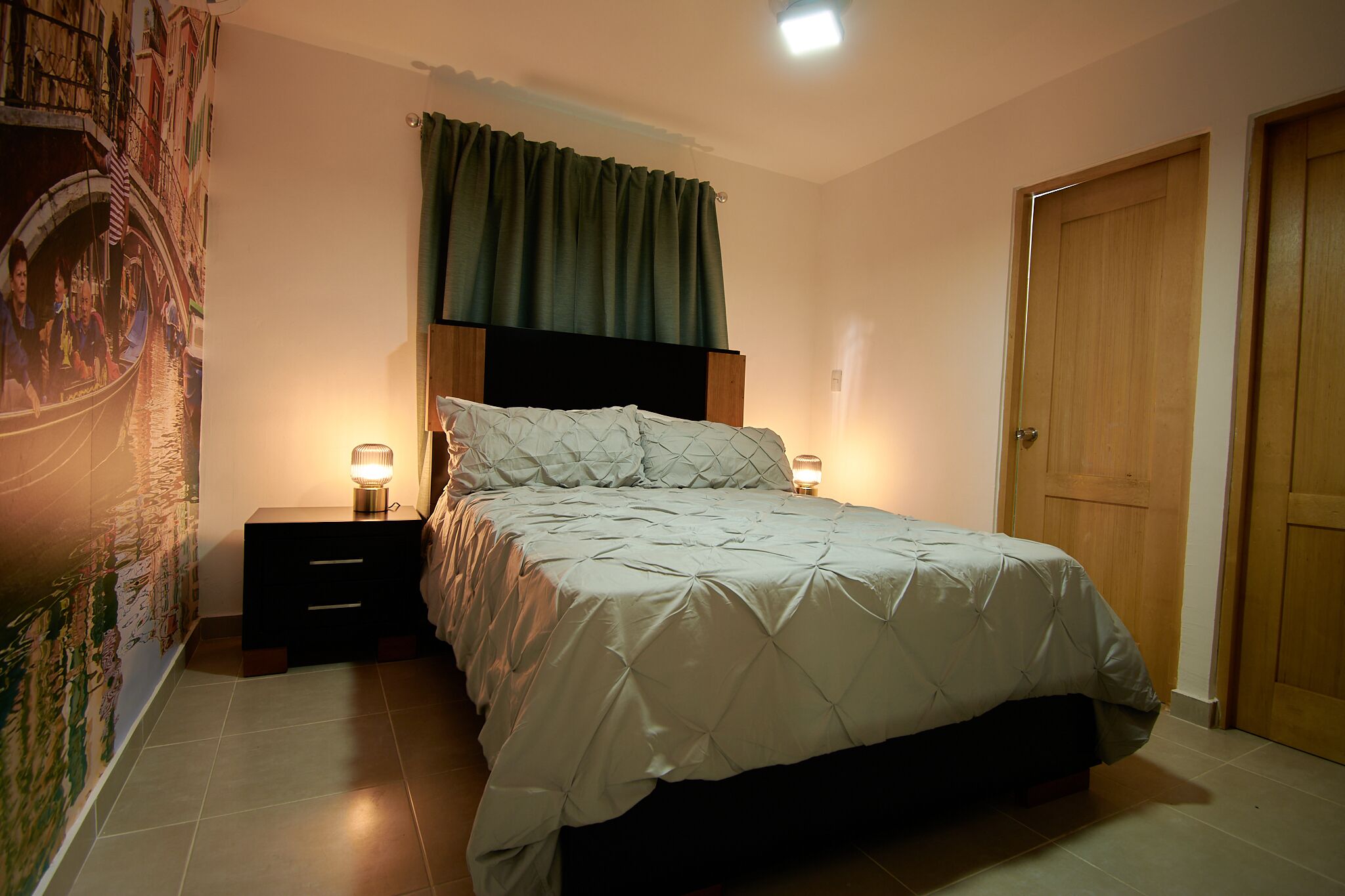Ein Schlafzimmer mit einem Bett, zwei Nachttischen mit Lampen, einem grünen Vorhang und einer Tür.