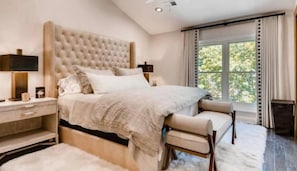 A modern bedroom with a large bed, bedside tables, a nightstand, and a view of greenery through the window.