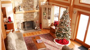 A cozy living room with a stone fireplace, wooden beams, a Christmas tree, and a large window.