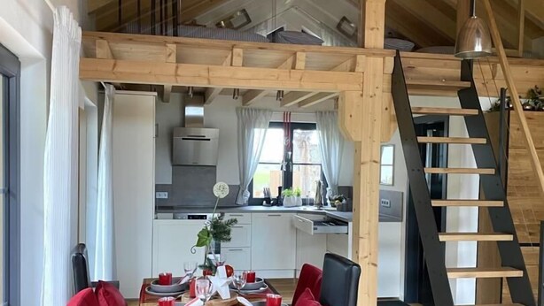 A dining area with a wooden table and red chairs, a kitchen in the background, and a wooden staircase. - Vacation cottage Schorenmoos-Lodge (Dietmannsried)