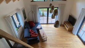 A spacious loft-style room with a wooden staircase, a blue sofa with red cushions, a wooden coffee table, and a TV on a wooden stand. - Vacation cottage Schorenmoos-Lodge (Dietmannsried)