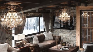 Un salon chaleureux avec un mur en pierre, une grande fenêtre donnant sur les arbres et deux lustres.