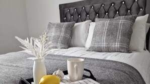 Un lit bien fait, recouvert d’un plaid rayé gris et blanc, un vase blanc contenant des fleurs séchées, un fruit jaune et une tasse blanche posés sur un plateau.