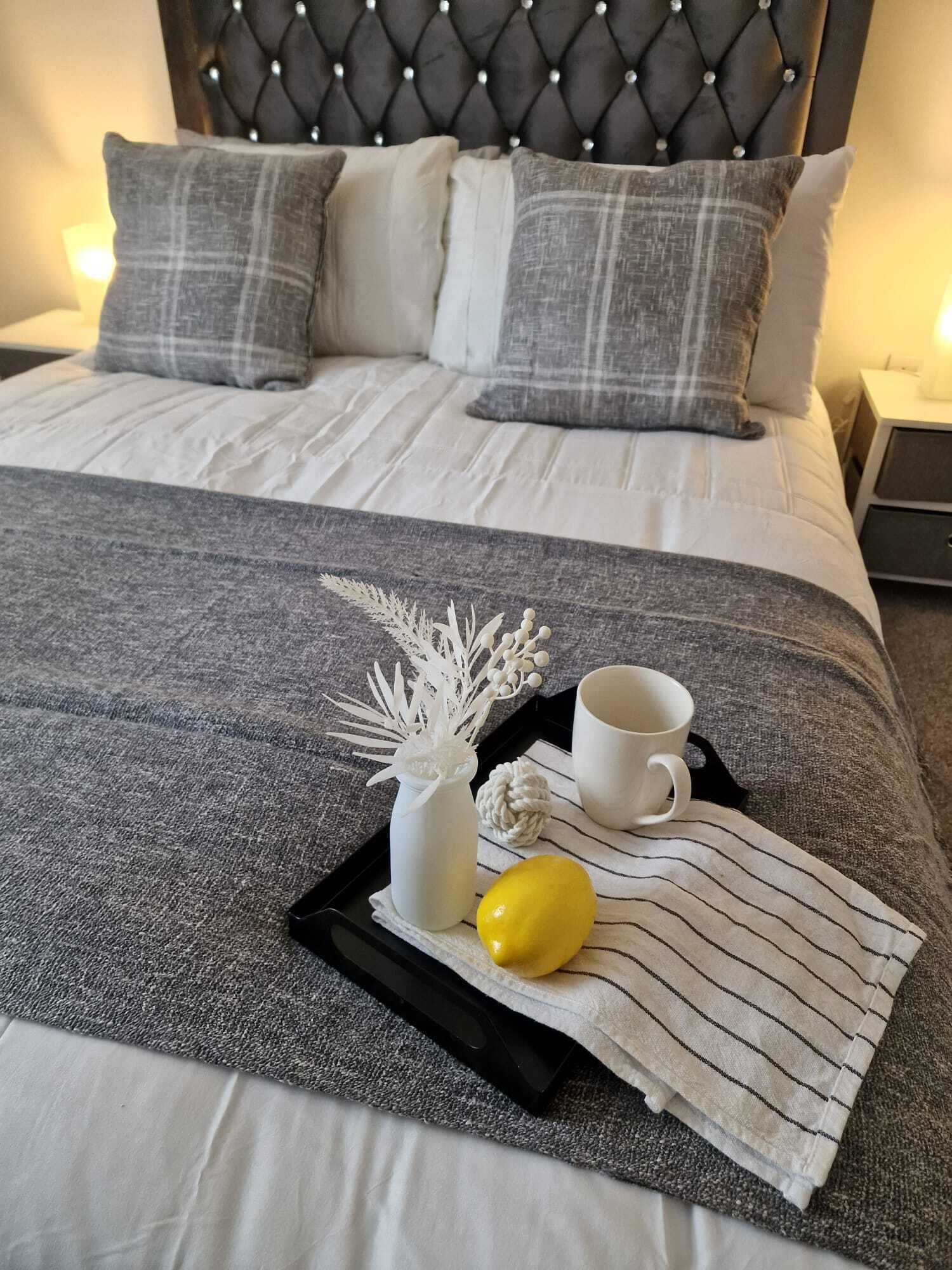 Un lit bien fait, recouvert d’une couverture à rayures grises et blanches, avec deux oreillers gris, un vase blanc contenant des branches décoratives, un citron et une tasse blanche posés sur un plateau.