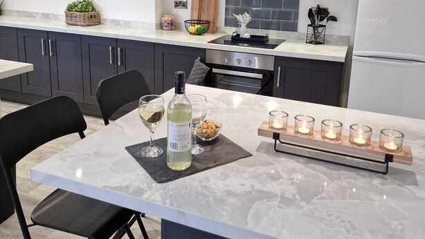 Une cuisine moderne avec des meubles de couleur sombre, un plan de travail blanc, un îlot central doté de chaises et un four encastré.