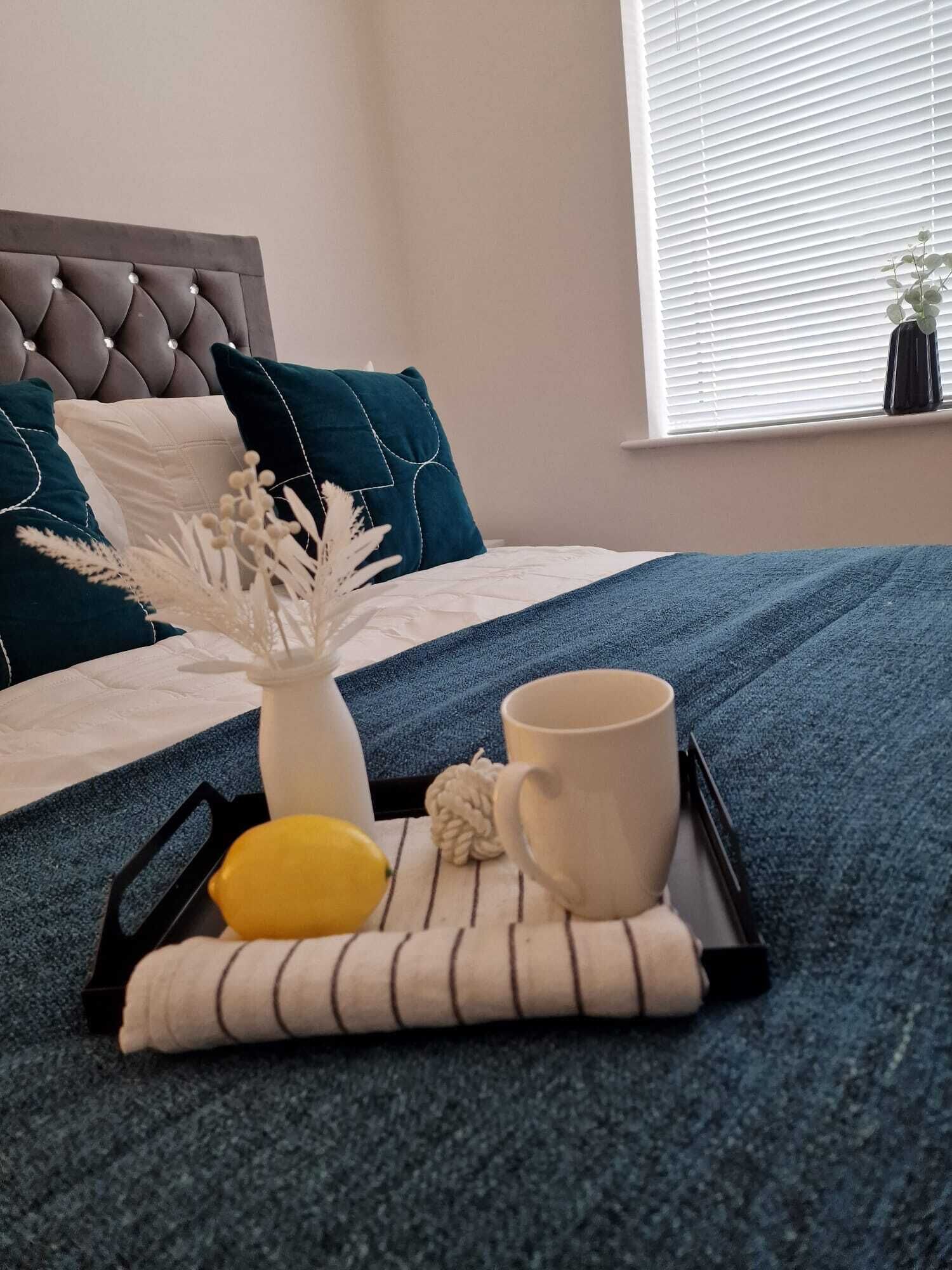 Una cama bien hecha con un edredón azul, un jarrón blanco con flores secas, un limón amarillo y una taza blanca sobre una bandeja.