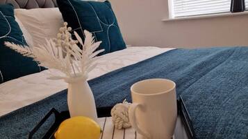 Una cama bien hecha con un edredón azul, un jarrón blanco con flores secas, un limón amarillo y una taza blanca sobre una bandeja.