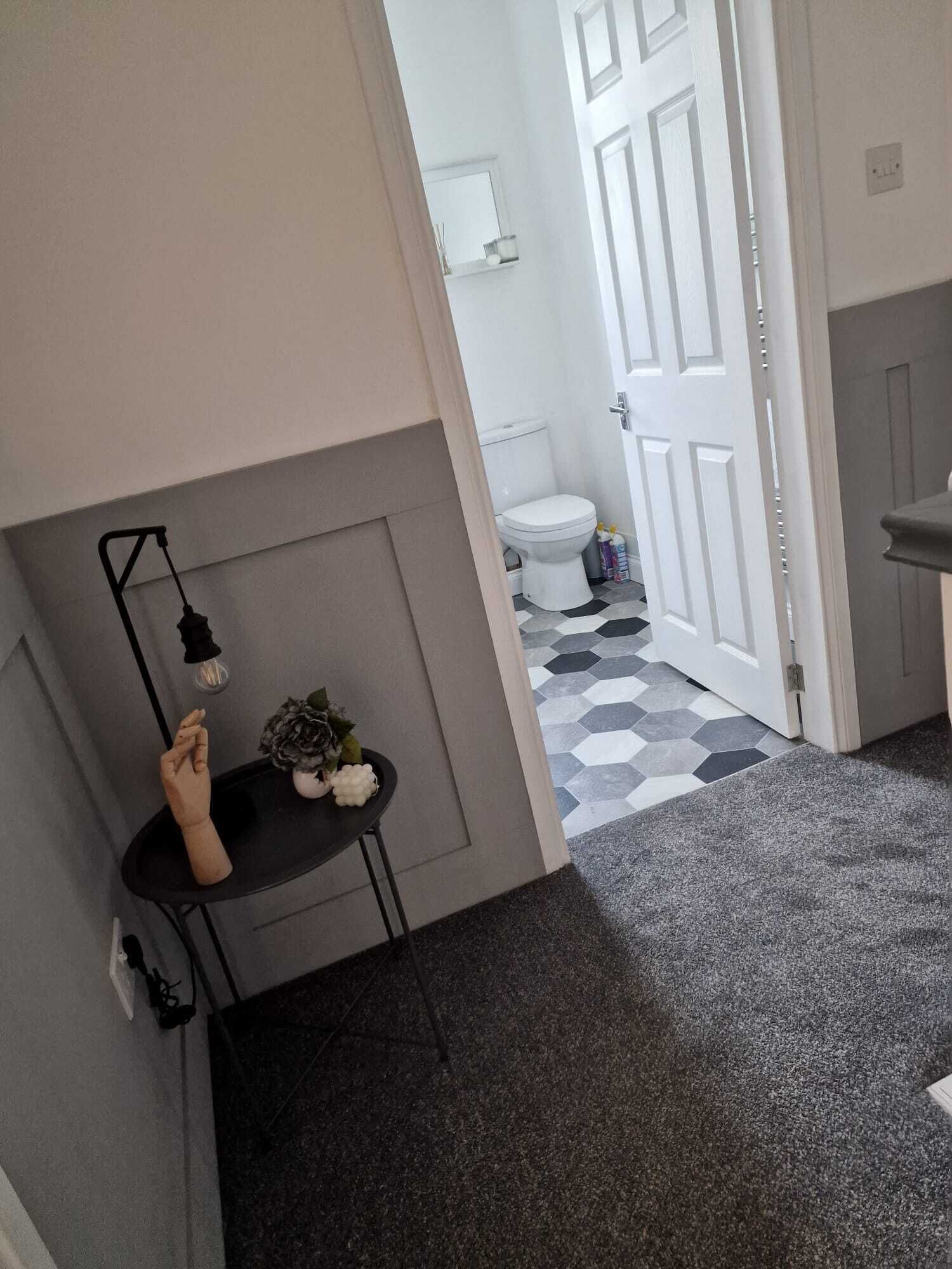 A narrow hallway with a black table and a potted plant, leading to a bathroom with a white toilet and a hexagonal tiled floor.
