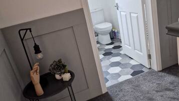 A narrow hallway with a black table and a potted plant, leading to a bathroom with a white toilet and a hexagonal tiled floor.