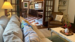A cozy living room with a sofa, armchair, and a coffee table. There is a checkered floor, a dining area in the background, and a birdcage on a shelf.