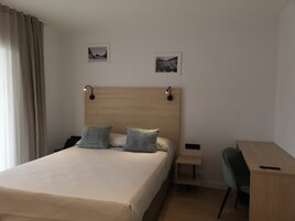 Quarto de hotel com uma cama grande, duas almofadas, uma mesa de cabeceira, uma escrivaninha e duas fotografias emolduradas na parede.