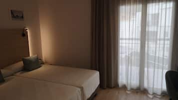 Quarto de hotel com duas camas, cabeceira, luminária fixada na parede e vista através de cortinas leves.