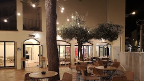 Un espace café en plein air avec des tables rondes et des chaises en osier, entouré d’arbres et illuminé de guirlandes lumineuses.