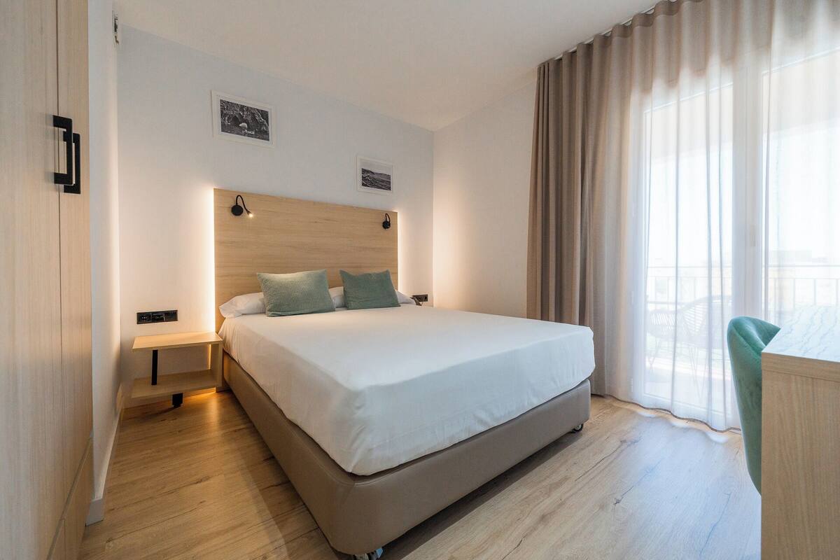 Een hotelkamer met een groot bed, nachtkastje, wandlamp en een raam met gordijnen.