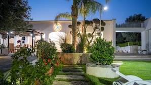 A modern outdoor area with a stone pathway, palm trees, and a building with a circular light fixture. - Villa Machri - Con Piscina Privata E Ampio Giardino Vicina Al Mare (Alessano)