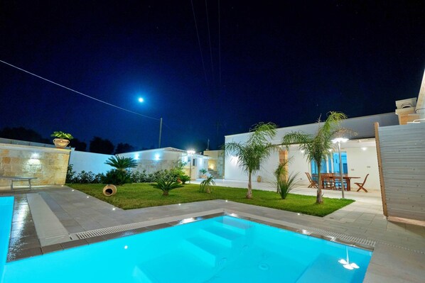 A swimming pool with a tiled deck, surrounded by a grassy area with palm trees and a patio with outdoor furniture.
