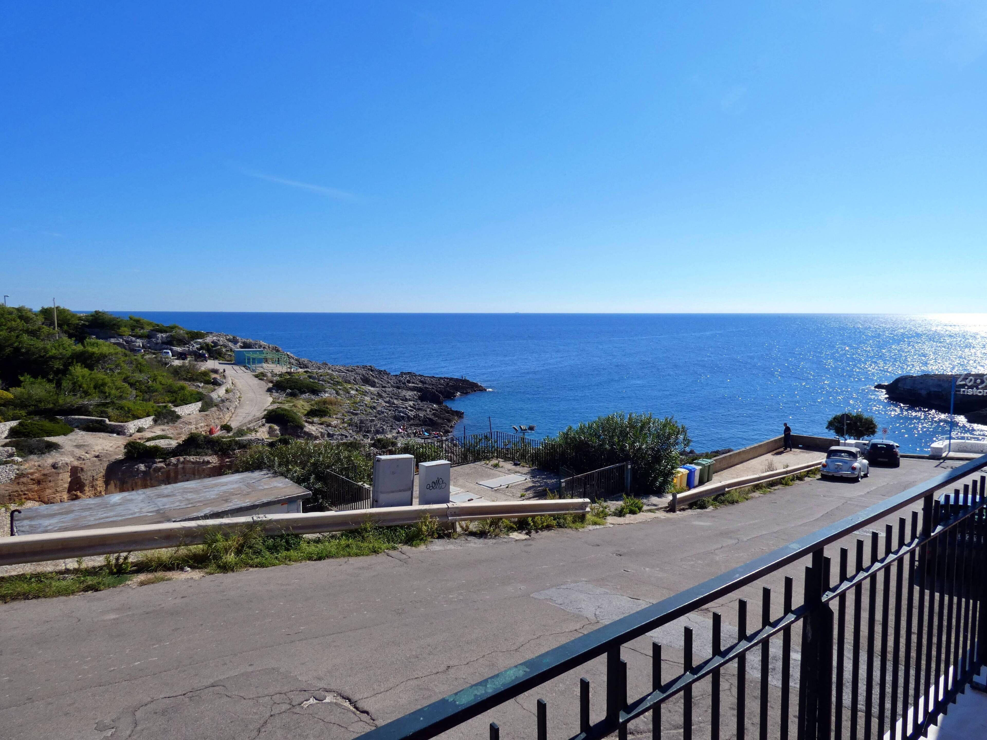 A coastal road with a view of the sea, a rocky area with vegetation, and parked cars.